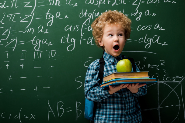 Überraschtes Kind hält Bücher und Apfel neben Kreidetafel mit mathematischen Formeln  - Foto, Bild