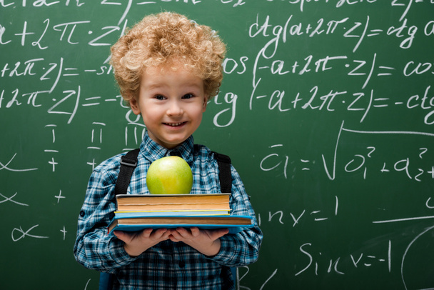 curly kid holding books and apple near chalkboard with mathematical formulas  - Fotoğraf, Görsel