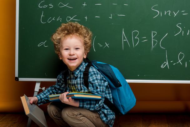 glückliches Kind lacht, während es Bücher mit mathematischen Formeln in der Nähe der Tafel hält  - Foto, Bild