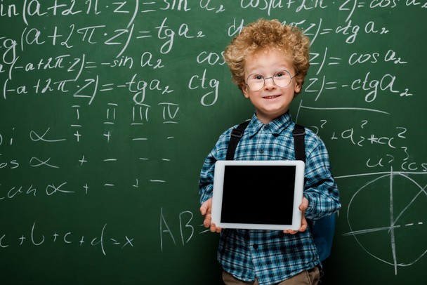 enfant heureux et intelligent dans des lunettes tenant tablette numérique avec écran blanc près de tableau noir avec des formules mathématiques
  - Photo, image