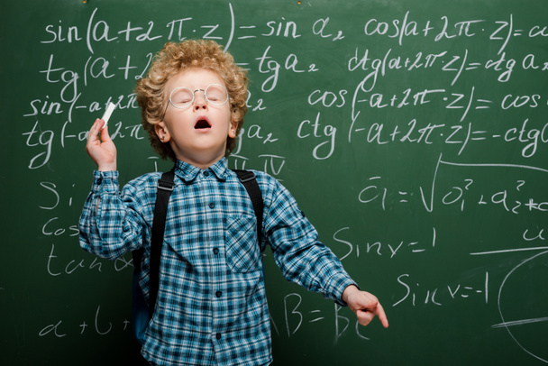 σγουρό παιδί σε γυαλιά φτέρνισμα κοντά σε μαυροπίνακα με μαθηματικούς τύπους  - Φωτογραφία, εικόνα