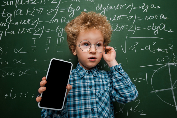 niño inteligente en gafas con teléfono inteligente con pantalla en blanco cerca de pizarra con fórmulas matemáticas
  - Foto, imagen