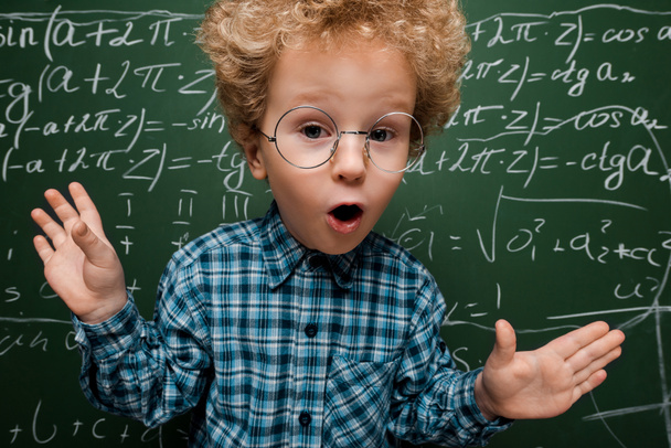 έκπληκτος παιδί σε γυαλιά κοιτάζοντας κάμερα κοντά σε πίνακα με μαθηματικούς τύπους  - Φωτογραφία, εικόνα