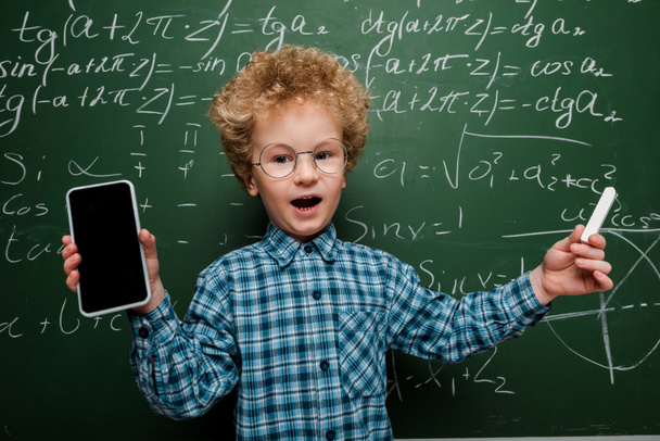 niedliches und intelligentes Kind, das ein Smartphone mit leerem Bildschirm in der Hand hält und Kreide neben eine Tafel mit mathematischen Formeln hält  - Foto, Bild