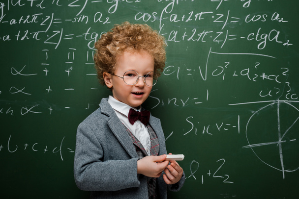 niño lindo e inteligente en traje con corbata de lazo sosteniendo tiza cerca de pizarra con fórmulas matemáticas
  - Foto, Imagen