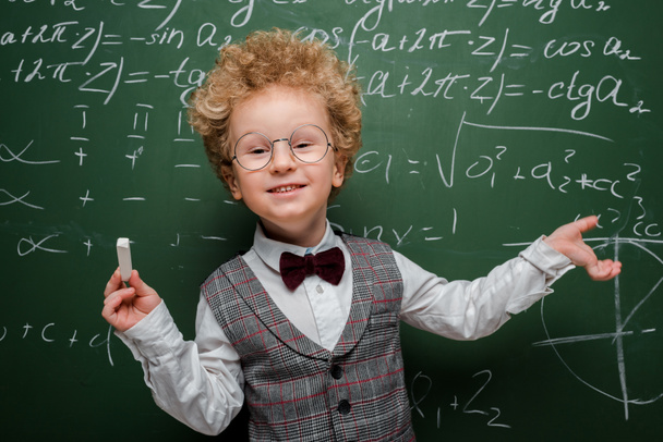 Lächelndes Kind in Anzug und Fliege, Kreide in der Hand und auf Tafel mit mathematischen Formeln zeigend  - Foto, Bild