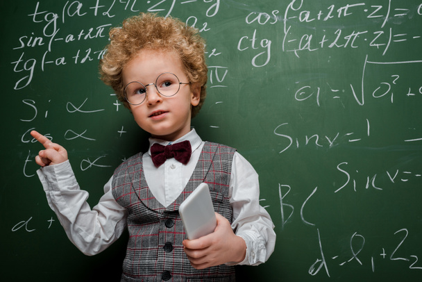 Smart Kind in Anzug und Fliege hält Smartphone in der Hand und zeigt mit dem Finger in die Nähe einer Tafel mit mathematischen Formeln  - Foto, Bild