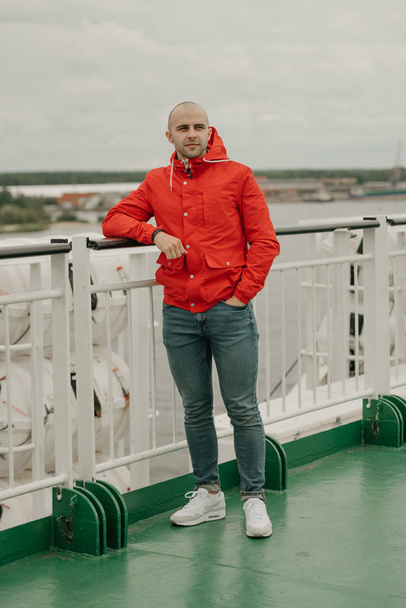 Calvo uomo brutale con stoppie in giacca rossa, jeans blu e scarpe da ginnastica bianche sulla nave godendo la sua crociera nel pomeriggio nuvoloso
 - Foto, immagini