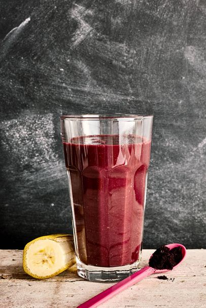 Single tall glass of exotic acai berry smoothie in between a slice of banana and plastic spoon over marble background - Fotoğraf, Görsel