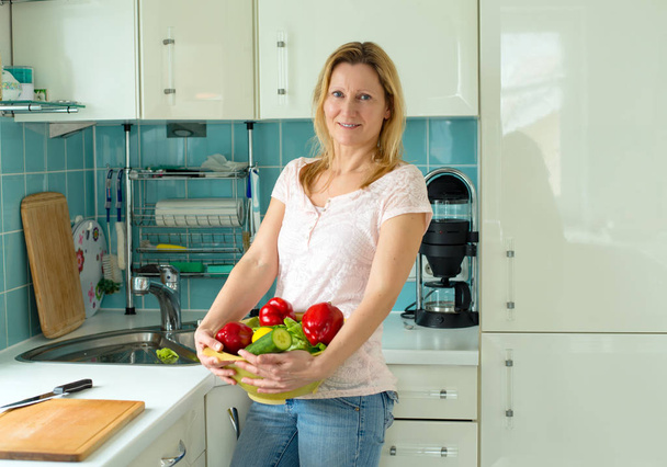 blond woman standing in the kitchen with vegetables in a bowl - Foto, Imagen