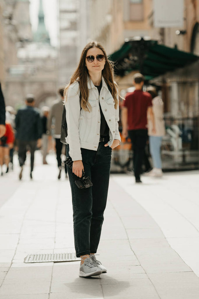Atraktivní mladá žena blogger v bílé džínové bundě, černé džíny, šedé tenisky a sluneční brýle drží stativ s chytrým telefonem pózuje v centru starověké evropské ulice plné lidí - Fotografie, Obrázek
