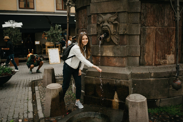 Een prachtige jonge vrouwelijke blogger in een witte jeans jas, zwarte jeans, grijze sneakers, zonnebril, en rugzak poseert en verheugde zich over de straal water in de buurt van de oude put op het centrum van het Europese plein   - Foto, afbeelding