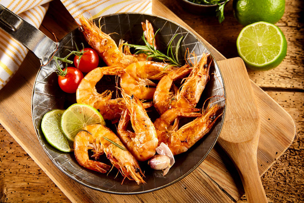 Delicious spicy grilled prawns with trimmings including fresh lime, garlic, cherry tomatoes and rosemary served in an old frying pan in a rustic country kitchen or restaurant - Fotografie, Obrázek