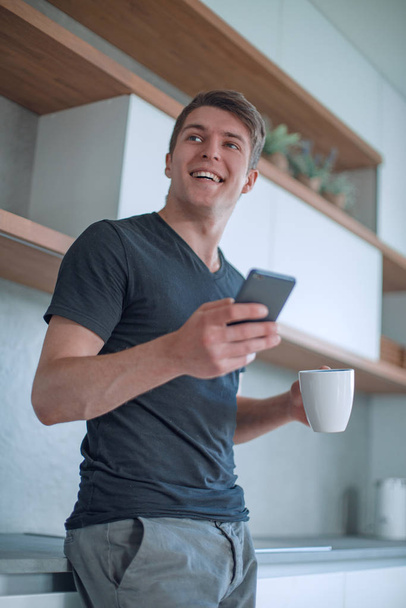 handsome guy with a Cup of coffee and a smartphone standing in the kitchen - Foto, imagen