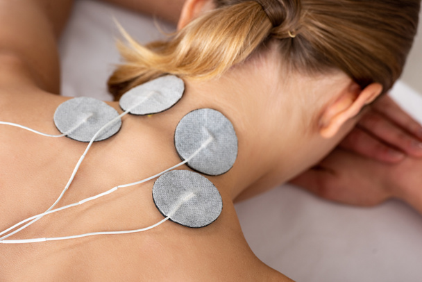 Visão traseira da mulher com eletrodos de eletroestimulação no pescoço no sofá de massagem isolado no cinza
 - Foto, Imagem