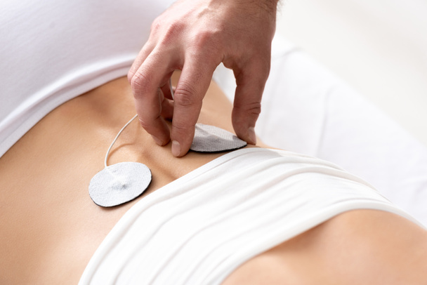 Ausgeschnittene Ansicht des Therapeuten, der die Elektrode auf dem Rücken des Patienten während der Elektrodenbehandlung auf grauem Hintergrund einstellt - Foto, Bild