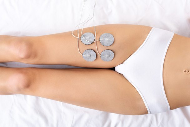 Top view of young woman in panties lying on massage couch during electrode treatment of leg   - Photo, Image