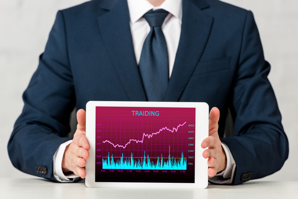 cropped view of businessman in suit holding digital tablet with trading courses on screen on white  - Photo, Image