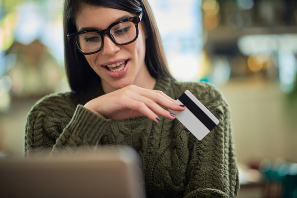 Attractive caucasian brunette in sweater sitting in cafe, typing on keyboard and holding credit card. - Photo, Image