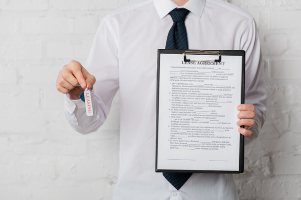 vista recortada de agente inmobiliario en traje con llave y portapapeles con contrato de arrendamiento en blanco
  - Foto, imagen