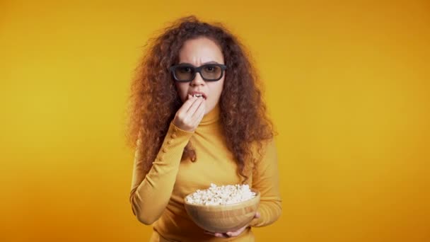 Young woman in 3d glasses watching movie and eating popcorn on yellow studio background.  - Filmagem, Vídeo