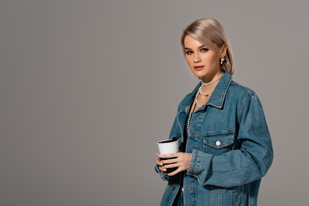 attractive woman in denim jacket holding insulated mug and looking at camera isolated on grey - Fotó, kép