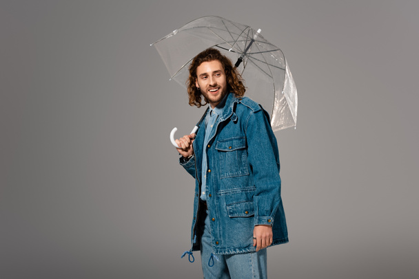hombre guapo y sonriente en chaqueta de mezclilla sosteniendo paraguas aislado en gris
 - Foto, Imagen