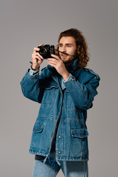 pohledný a stylový muž v džínové bundě a džínách fotografování izolované na šedé - Fotografie, Obrázek