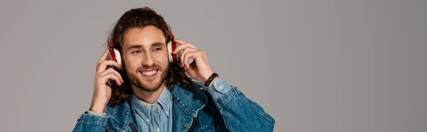 panoramic shot of smiling man in denim jacket listening to music with headphones isolated on grey - Fotoğraf, Görsel