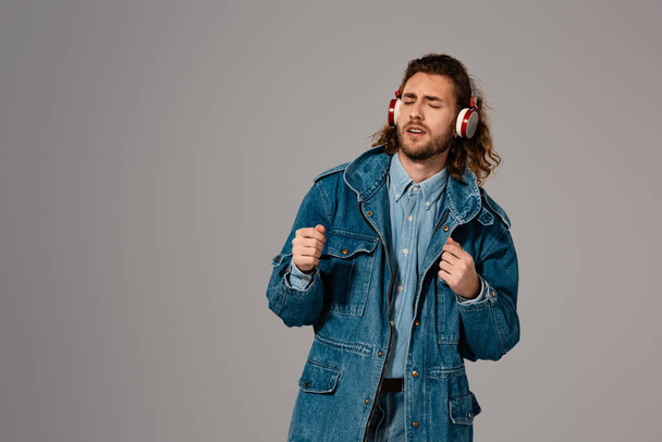 przystojny i stylowy mężczyzna w dżinsowej kurtce słuchający muzyki ze słuchawkami odizolowanymi na szaro - Zdjęcie, obraz