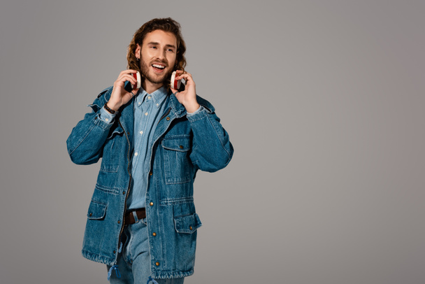 handsome and smiling man in denim jacket and jeans with headphones isolated on grey - Photo, Image