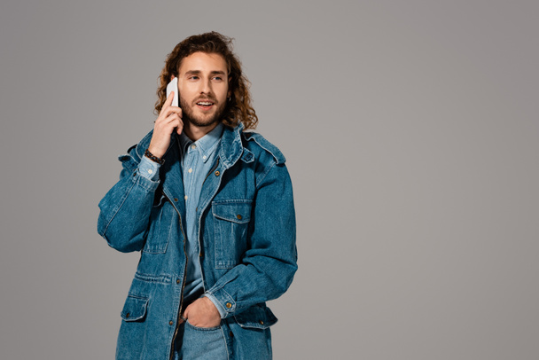 handsome and stylish man in denim jacket talking on smartphone isolated on grey - Foto, afbeelding
