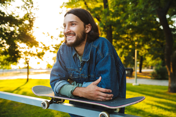 Smiling positive optimistic young bearded man with skateboard standing in nature park. - Foto, afbeelding