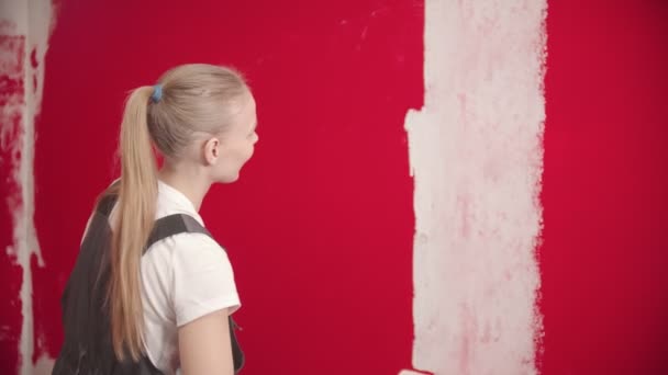 Mujer en overol está pintando una pared roja con un rodillo blanco
 - Metraje, vídeo