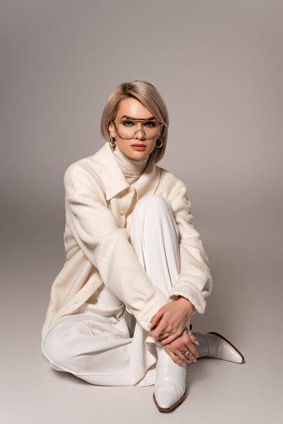 attractive woman in white coat and trousers looking at camera on grey background  - Fotografie, Obrázek