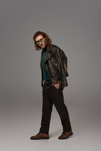 handsome man in leather jacket with hand in pocket looking at camera on grey background  - Foto, afbeelding