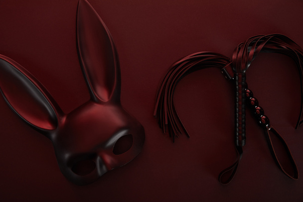 top view of rabbit mask and flogging whip in dark lighting on red background - Photo, Image