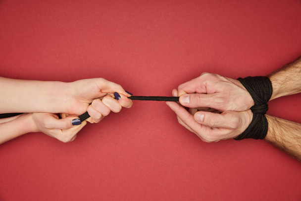 cropped view of woman holding rope on tied male hands isolated on red - Фото, изображение
