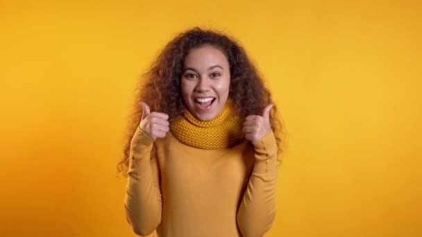 Winner. Success. Positive girl making thumbs up sign over yellow background and smiles to camera. Body language. Young mixed race curly woman - 映像、動画