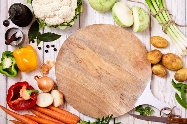 Round empty cutting board surrounded by fresh raw vegetables as carrots, potatoes, onion, pepper, cauliflower, kohlrabi, herbs and spices on a rustic wooden table, close up from above - Photo, image