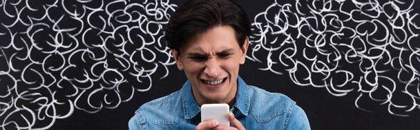 Panoramaaufnahme eines besorgten jungen Mannes mit Smartphone, mit Dampf aus den Ohren, der auf Kreidetafel gezeichnet wird - Foto, Bild