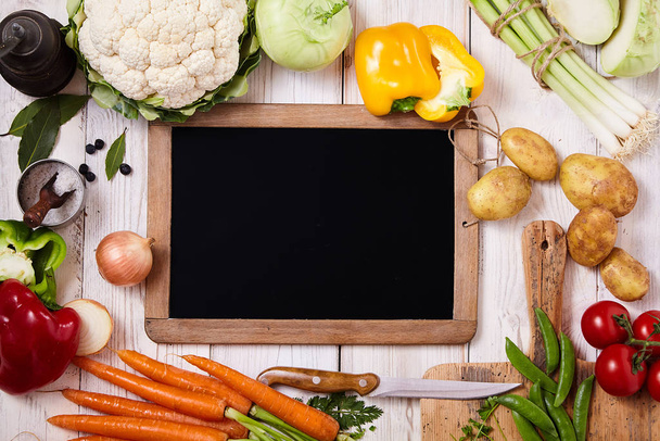 Blank rectangular chalkboard with wooden frame surrounded by fresh vegetables as onion, carrots, pepper, carrots, tomatoes, cauliflower, herbs and spices on a rustic table, close up from above - Photo, image