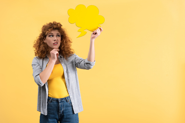 mooie sceptische vrouw op zoek naar lege spraak bel in de vorm van een wolk, geïsoleerd op geel  - Foto, afbeelding
