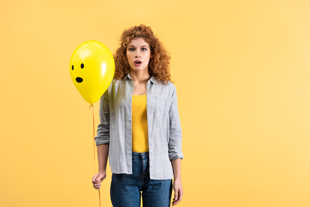 surprised redhead girl holding balloon with shocked face, isolated on yellow - Photo, Image
