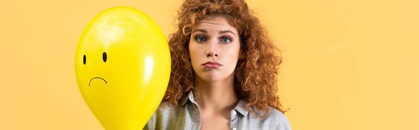 panoramic shot of upset redhead woman holding balloon with sad face, isolated on yellow - 写真・画像