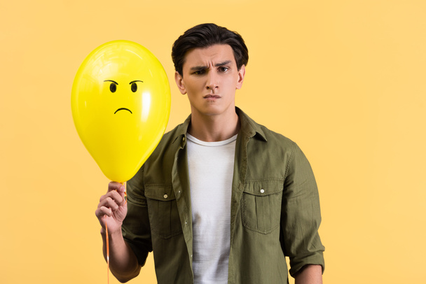 σκεπτικιστής νεαρός άνδρας που κρατά θυμωμένο μπαλόνι, απομονωμένος στο κίτρινο - Φωτογραφία, εικόνα