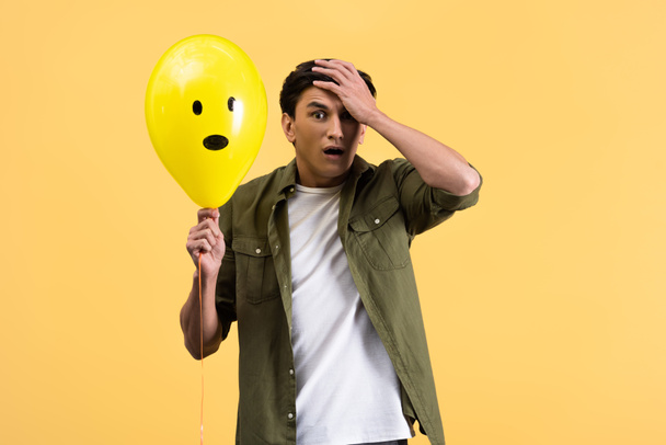 Ανησυχημένος νεαρός κρατώντας σοκαρισμένο μπαλόνι, απομονωμένος στο κίτρινο - Φωτογραφία, εικόνα