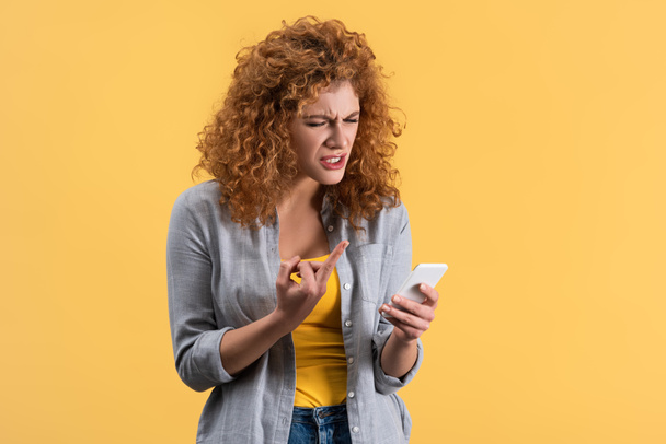 stressed redhead woman showing middle finger to smartphone, isolated on yellow - Zdjęcie, obraz
