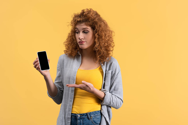 mujer pelirroja escéptica mostrando teléfono inteligente con pantalla en blanco, aislado en amarillo
 - Foto, Imagen