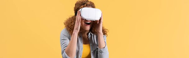 panoramiczne ujęcie podnieconej rudej kobiety korzystającej z zestawu słuchawkowego wirtualnej rzeczywistości, odizolowanej na żółto - Zdjęcie, obraz